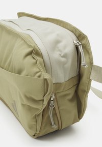 Bolsa de viaje rectangular color caqui con una parte superior de suave gris claro, múltiples cremalleras y tela texturizada. Cuenta con asas laterales y un diseño minimalista.