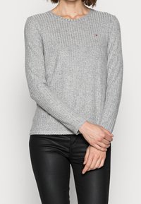 Kvinna klädd i en grå ribbad långärmad tröja och svarta läderbyxor, stående med händerna knäppta framför sig mot en enfärgad bakgrund.