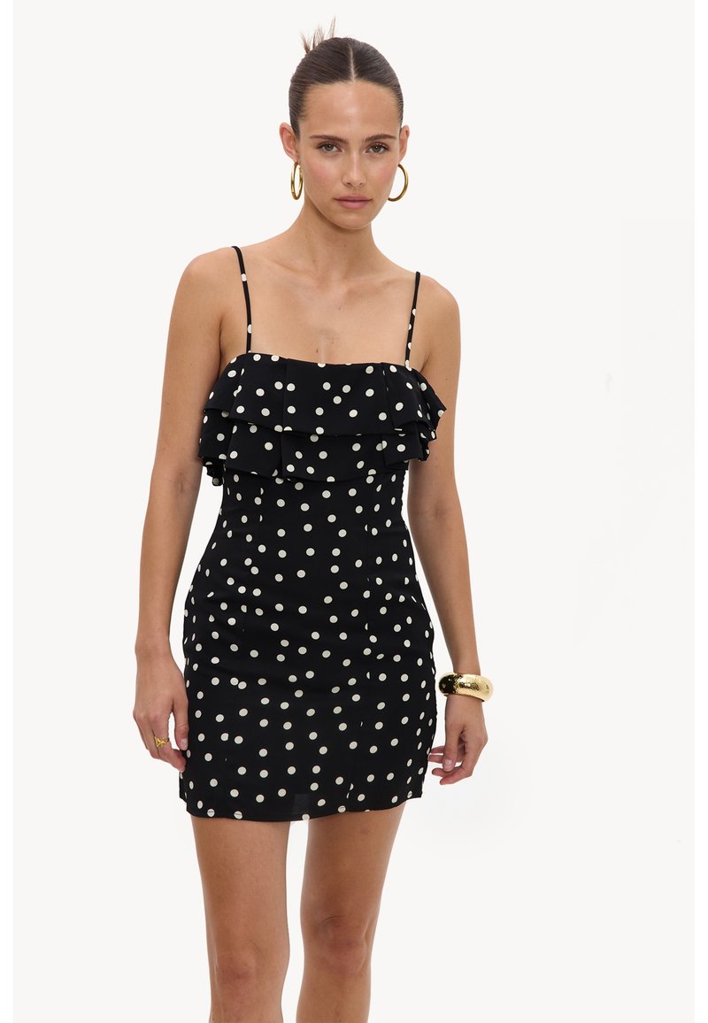 Femme portant une mini robe noire sans manches à pois blancs avec un détail volanté, accessoirisée de boucles d'oreilles créoles dorées et d'un bracelet en or.