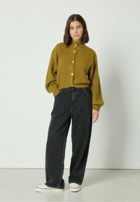 Olivno zelena pletena jopica z visokim ovratnikom in zapiranjem s gumbi, v kombinaciji z širokimi črnimi jeansa in belimi športnimi copati.