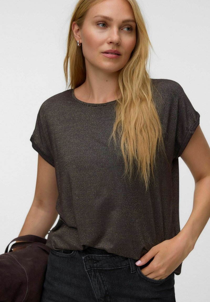 Femme blonde portant un haut ample sombre à manches courtes et un jean foncé, tenant une veste sombre dans une main et l'autre main dans la poche.