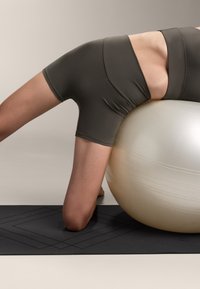 Szare spodenki sportowe zakrywają dolną część ciała. Osoba opiera się o jasnokolorową piłkę do ćwiczeń, opierając się na czarnej macie do ćwiczeń z geometrycznym wzorem.