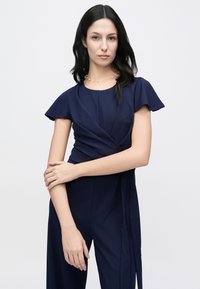 Mono azul marino con mangas cortas con volantes, diseño cruzado y pantalones anchos. Textura de tela suave con un brillo sutil. Cuenta con un detalle de cintura anudada.