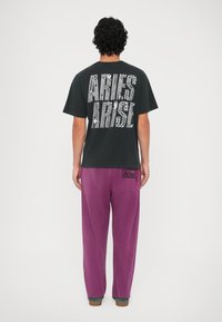 Fekete póló nagy, fehér kristályokkal díszített "ARIES ARISE" felirattal a hátsó részén, lazán illeszkedő lila nadrággal, amelyen logó folt található.