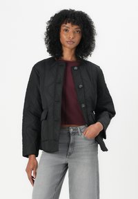Vrouw met krullend haar, gekleed in een zwarte gewatteerde jas, een bordeaux crop top en lichtgrijze spijkerbroek, staand met haar hand in haar zak tegen een witte achtergrond.
