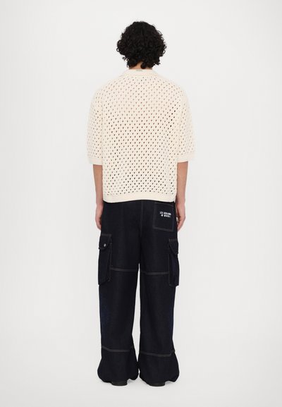 Pull en maille crème à manches courtes avec un motif en mailles ouvertes, associé à un pantalon cargo en denim foncé à jambes larges doté de plusieurs poches et d'une étiquette logo au dos.