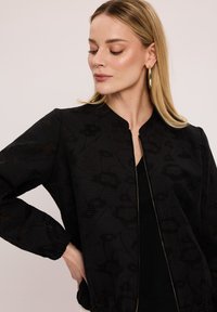 Czarna kurtka bomberka z fakturowanym wzorem, z zapięciem na zamek z przodu i elastycznymi mankietami. Posiada kołnierz mandarynki oraz luźny krój.