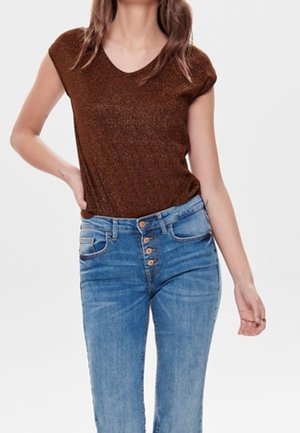 Vrouw die een bruin kortärmelig topje en een high-waisted blauwe spijkerbroek met zichtbare knopen draagt, staand tegen een effen achtergrond.