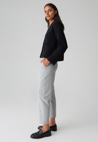 Pull côtelé noir avec une poche plaquée, associé à un pantalon gris clair bien taillé et des mocassins noirs, présenté sur un fond neutre.