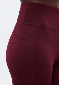 Gros plan d'une personne portant un legging bordeaux taille haute avec une poche latérale et des détails de couture visibles.