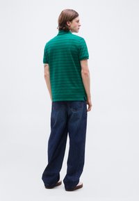 Jeune homme aux cheveux mi-longs portant un polo rayé vert, un jean bleu foncé ample et des chaussures marron, vu de profil.