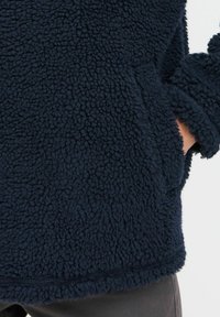 Marineblå fleece-genser med en teksturert, fluffy overflate, med kengurulomme og brede ermer, nederst sydd for fleksibilitet.
