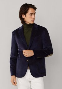 Blazer de pana en color navy con solapa muesca, dos bolsillos frontales y parches de codo en color beige. Combinado con un cuello de tortuga verde oscuro y pantalones blancos.