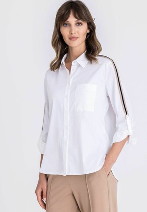 Femme portant une chemise blanche à boutons avec des rayures noires et beiges sur les manches et un pantalon beige, debout avec une main dans la poche.