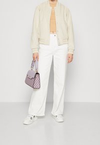 Blouson aviateur crème avec poignets rayés, crop top orange clair, pantalon large blanc et sac à main à carreaux violet. Des baskets blanches complètent le look.