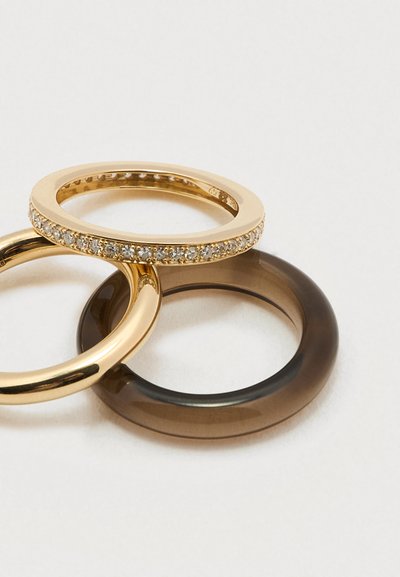 Három egymásra helyezett gyűrű: egy aranysáv apró, tiszta kövekkel, egy sima aranysáv, és egy áttetsző sötétbarna sáv világos háttéren.