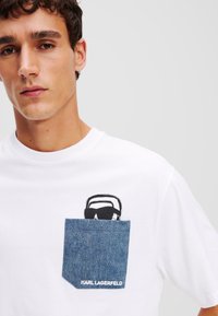 Balta marškinėlių su džinsiniu kišeniu, ant kurio pavaizduoti juodai balti saulės akiniai ir šiuolaikinio šrifto užrašas "KARL LAGERFELD".