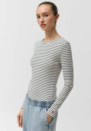 Kobieta w białej koszuli z długim rękawem w cienkie czarne paski z małym czerwonym sercem na mankiecie, w połączeniu z jasnoniebieskimi dżinsami.