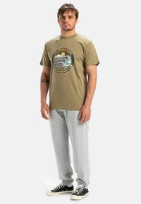 Olivgrünes Baumwoll-T-Shirt mit kreisförmigem Grafikdesign auf der Vorderseite, kombiniert mit hellgrauen Sweatpants und schwarzen Turnschuhen.