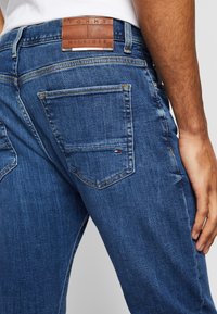Närbild av en person som bär blå Tommy Hilfiger-jeans, som visar bakfickan med en liten logotyp och lädermärke ovanför midjebandet.