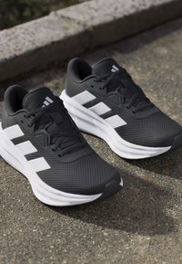 Chaussures de course Adidas noires et blanches avec trois bandes sur l'asphalte à côté d'un trottoir, en angle pour montrer la vue de dessus et de profil.
