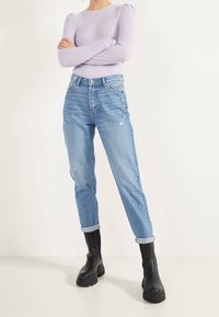 Persona con suéter de canalé color púrpura claro, jeans azul claro con puños y pequeñas marcas de desgaste, y botas negras gruesas con plataforma, de pie con los brazos cruzados.