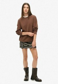 Felpa marrone con colletto bianco, minigonna mimetica e stivali di pelle neri fino al ginocchio. Modella in piedi con le braccia incrociate, frontale.