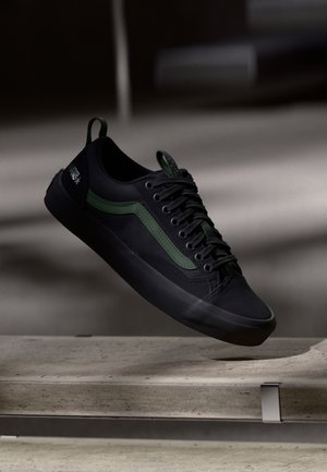 Zapatilla negra con acentos verdes, parte superior de tela, punta redonda, suela plana y diseño con cordones. Incluye un logo en el talón y una lengüeta texturizada.