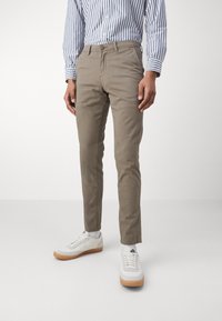 Chinos in cotone marrone chiaro con una vestibilità slim, dotati di chiusura a bottone e tasche laterali, abbinati a sneakers bianche. Camicia a righe blu e bianca.