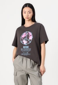 Mørkebrun t-shirt med en løs pasform, der har et pink og blåt grafisk design af en globus og teksten "NATURE VS INDUSTRY." 100% bomuldsstof, korte ærmer.
