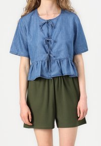 Top in denim azzurro chiaro, taglio cropped, con maniche corte e orlo peplum, dotato di tre lacci frontalmente. Indossato con pantaloni corti elasticizzati in vita di colore verde scuro.