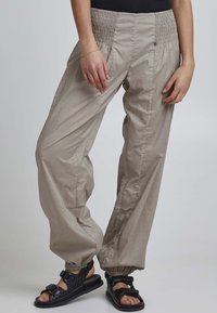 Light beige, lightweight pants with a smocked waistband; elastic cuffs; paired with black sandals featuring adjustable straps and textured design.