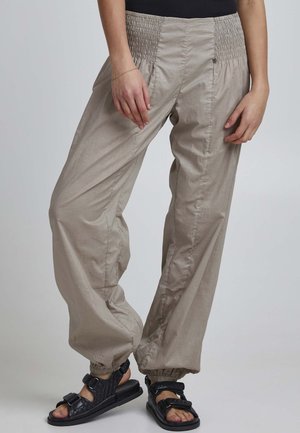 Light beige, lightweight pants with a smocked waistband; elastic cuffs; paired with black sandals featuring adjustable straps and textured design.