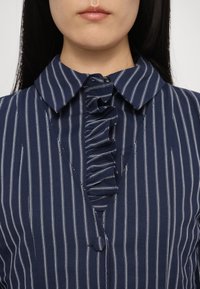 Femme portant un chemisier bleu marine à rayures blanches verticales avec un plastron à volants boutonné, montrant les détails du décolleté et du col.