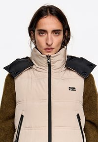 Gilet imbottito beige con collare nero a scatto, dotato di cerniere frontali e logo "Bimba y Lola". Tessuto esterno liscio; texture interna morbida.
