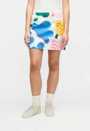 Γυναίκα που φοράει λευκή μίνι φούστα με μεγάλα μπλε, κίτρινα, ροζ και πράσινα λουλουδένια σχέδια, λευκές κάλτσες και μπεζ αθλητικά παπούτσια.