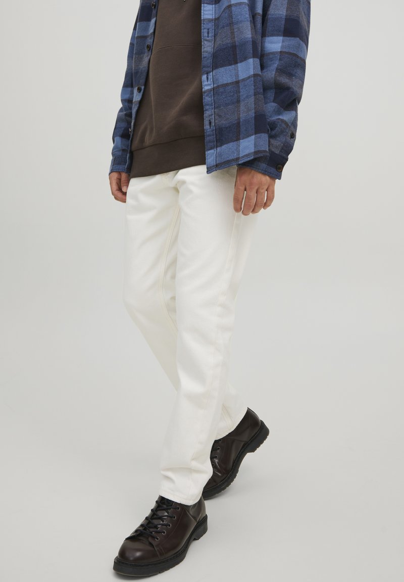 Pantalons blancs slim fit associés à un sweat à capuche marron et une chemise à carreaux bleue à manches longues, agrémentés de bottes noires à lacets.