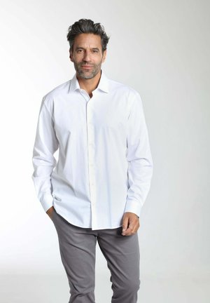 Homme aux cheveux poivre et sel et à la barbe, portant une chemise blanche à bouton et un pantalon gris, debout avec une main dans la poche.