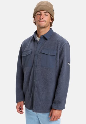 Fleecehemd in Marineblau mit Knopfleiste, zwei Brusttaschen und strukturiertem Stoff. Kombiniert mit hellblauen Jeans und einer beigen Strickmütze.