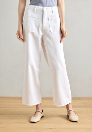 Kobieta ubrana w białe spodnie o szerokich nogawkach z dużymi przednimi kieszeniami oraz beżowe baleriny typu Mary Jane, stojąca na drewnianej podłodze.