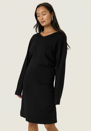 Frau in einem schwarzen, knielangen Kleid mit langen Ärmeln und einem subtilen Nahtdetail, trägt silberne Creolen und steht vor einem einfarbigen Hintergrund.