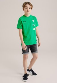 Zielona bawełniana koszulka z białymi grafikami, w tym palmą, falami i okrągłym kształtem; w komplecie z podwiniętymi dżinsowymi szortami i czarnymi sneakersami.