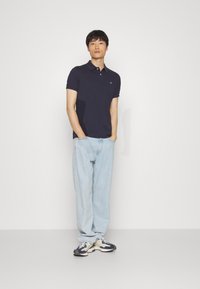 Marineblaues Polohemd mit Knopfleiste, getragen mit hellblauen, locker sitzenden Jeans und mehrfarbigen Sneakers vor einem einfarbigen Hintergrund.