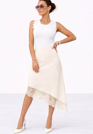 Femme portant un haut blanc sans manches et une jupe beige asymétrique avec un ourlet en dentelle, des talons hauts blancs, des lunettes de soleil et des bijoux, posant avec confiance.