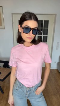 Noor naine, kes kannab sinaka tooniga päikeseprille, roosat Calvin Kleini T-särki ja hele siniseid teksapükse, seisab toas ühe käe taskus.