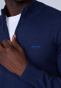 Pull zippé bleu marine avec un col haut. Fabriqué en tissu texturé et orné d'un petit logo d'animal bleu sur la poitrine. Manches ajustées.