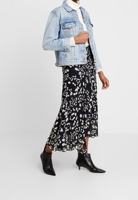 Veste en denim bleu clair avec fermeture à boutons, portée sur une jupe noire avec un motif abstrait blanc, accompagnée de bottines noires.