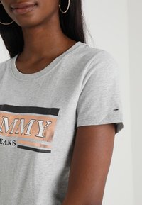 Camiseta de algodón gris con un logo impreso en negro y oro rosa. Manga corta con escote redondo y un pequeño detalle del logo en la manga.