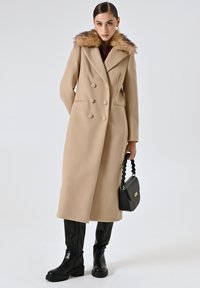 Cappotto di lana beige con colletto in pelliccia sintetica staccabile, design doppiopetto e maniche lunghe; abbinato a stivali neri con tacco e borsa.
