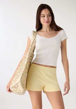 Top bianco a maniche corte con rifiniture in pizzo, abbinato a pantaloncini giallo chiaro. Tiene una borsa tote beige con motivo leopardato.